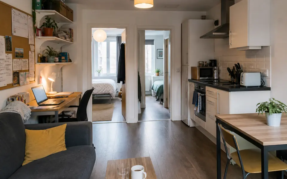 Interior of a shared student flat in London, showing a small living area with sofa and desk next to a compact kitchen, with bedroom doors leading off a hallway.